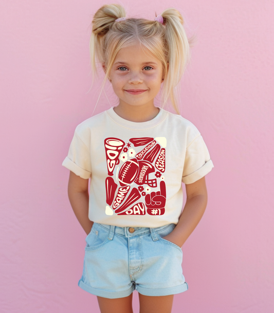 A toddler wearing a beige t-shirt with a red and black floral touchdown mascot design, paired with blue denim shorts, standing against a pink background.